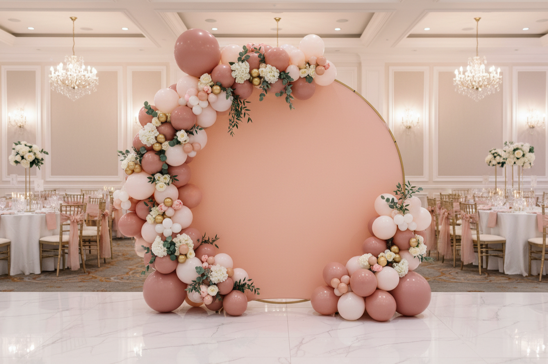 Elegant wedding backdrop featuring a blush pink round arch with a pastel balloon garland, white flowers, greenery, and gold accents in a grand ballroom
