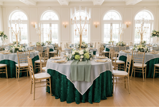 Elegant wedding reception with emerald green velvet tablecloths, white overlay linens, gold Chiavari chairs, tall gold candle centerpieces, and lush white floral arrangements in a bright ballroom