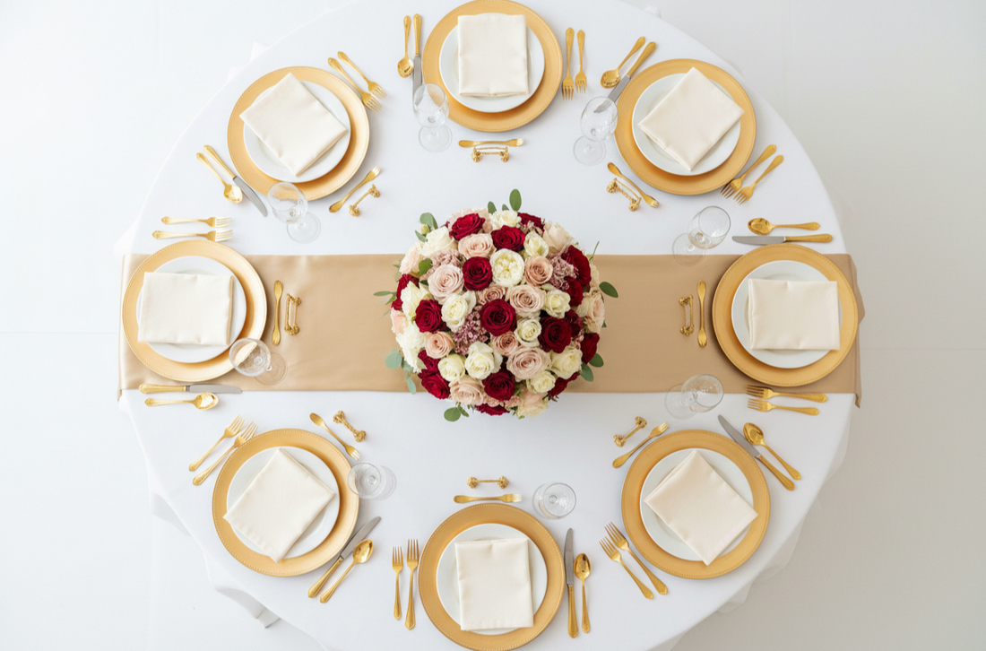 Top view of a round table with white linens, gold place settings, a champagne runner, and a red and blush floral centerpiece