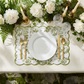 French Toile scalloped-edge placemats in olive green under a white plate with gold cutlery and a floral centerpiece.