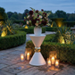 31" Tall Hourglass Plinth Pedestal Stand White outdoor garden set up with gold pillar candles, and a vase of mixed florals