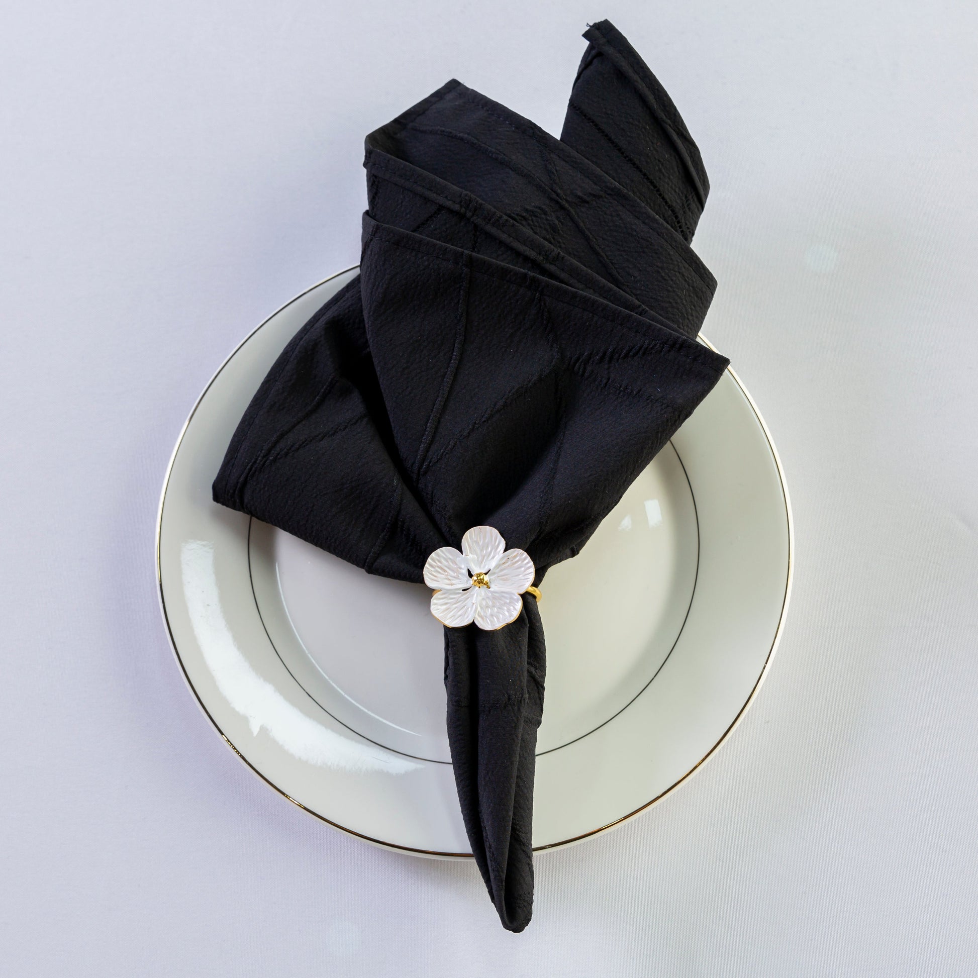 5 pcs/pk Embossed Geometric Jacquard Napkin 20"x20" Black fan fold with a white flower napkin ring on top a white dinner plate
