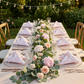 outdoor table setting with the French Toile 20"x20" Linen Napkin (5pc/pk) - Dusty Rose/Mauve in a triangle fold on glass charger plate and floral and greenery table runner
