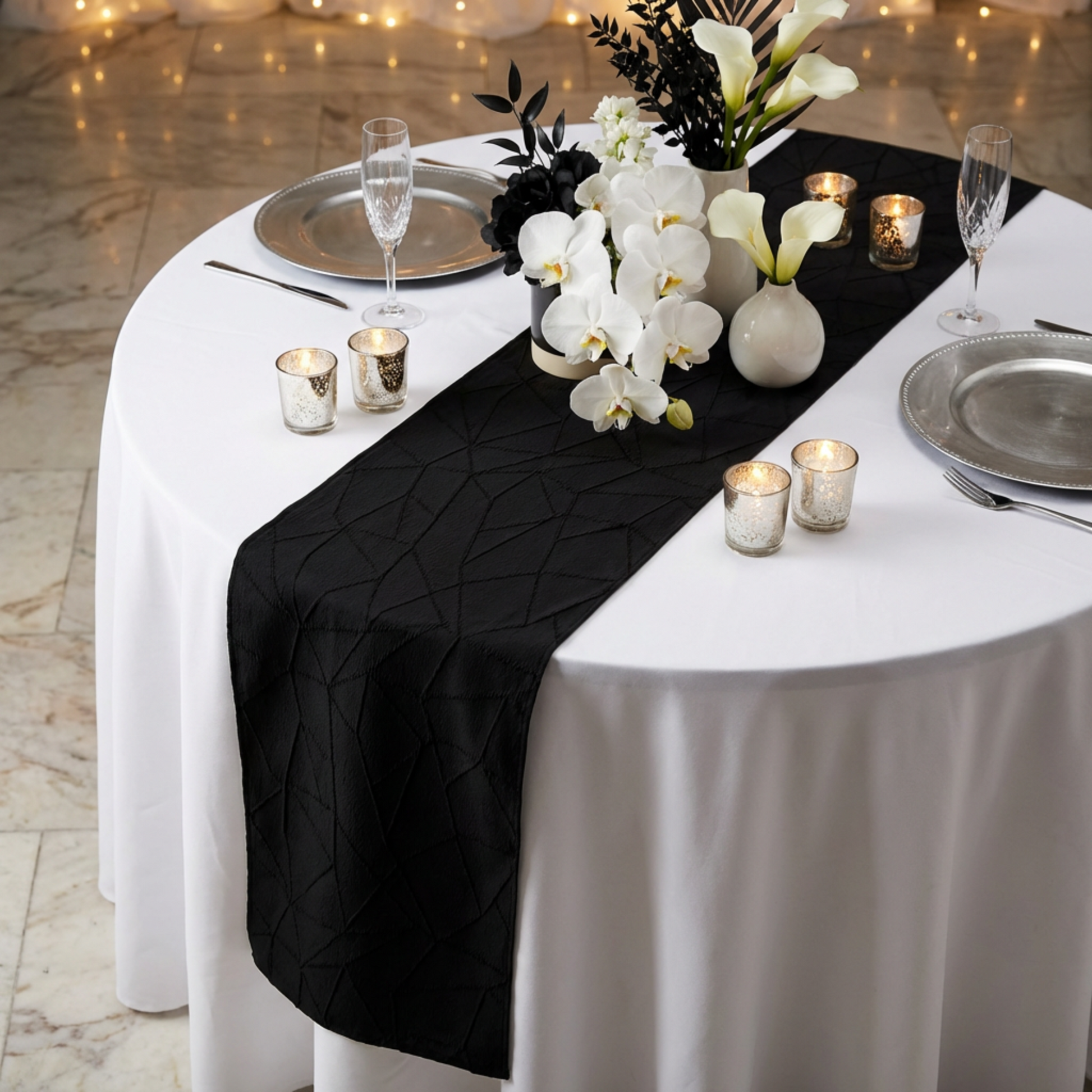 Embossed Geometric Jacquard Table Runner Black on top a round white tablecloth paired with silver charger plates, white and black flrals, silver tea light candle holders