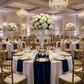 Glitter Velvet Table Runner Navy Blue on top a white tablecloth paired with gold charger plates, wine goblets and tall white floral centerpiece