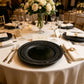 Black lace-embossed acrylic charger plate on a formal table setting with silverware and a rose centerpiece.