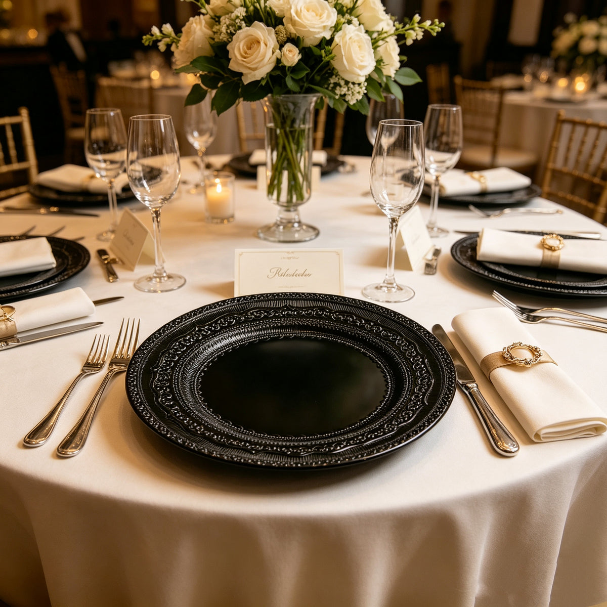 Black lace-embossed acrylic charger plate on a formal table setting with silverware and a rose centerpiece.