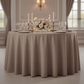 Premium Polyester 108" Round Tablecloth Taupe on a 60" round table with tapered candles, wine goblets, white charger plates, and gold silverware