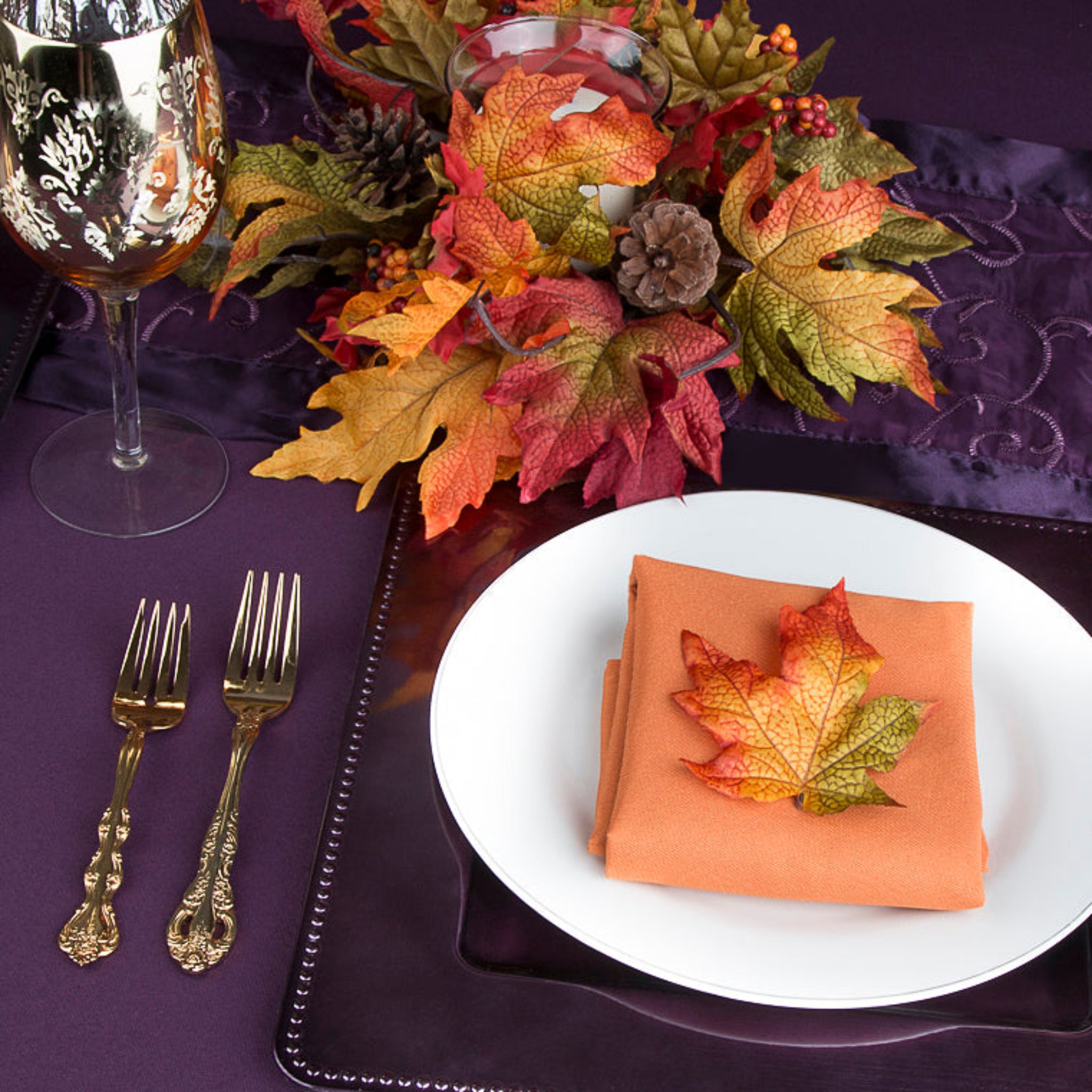 Burnt orange polyester napkin folded on a white plate amid autumn leaves in a purple table setting.