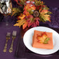 Burnt orange polyester napkin folded on a white plate amid autumn leaves in a purple table setting.