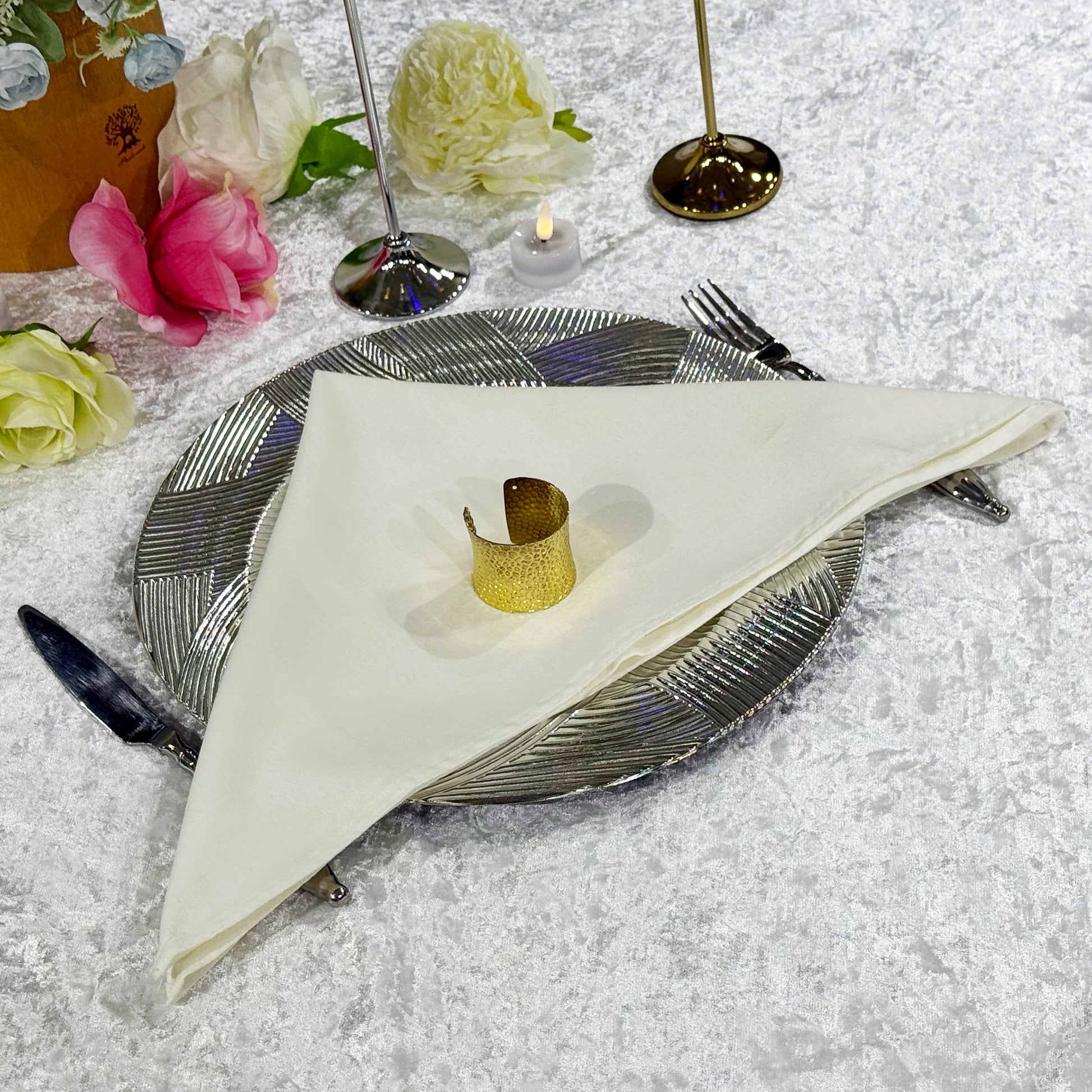 Premium Spun Polyester Napkin 20"x20" Ivory on top a silver charger plate with a gold napkin ring and silverware