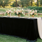 decorative outdoor tablescape of Velvet King 108"x156" Rectangular Tablecloth - Black setup with flowers, tapered candles, a cocktail glass, and chiffon table runner
