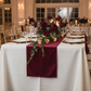reception table with the Velvet Table Runner Cabernet  on top a white tablecloth, gold charger plates, pillar candles and burgudy floral and greenery centerpiece