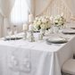 Vintage Veil Embroidery Table Runner White on top a white tablecloth with  pillar candles, white roses centepiece, LED backdrop curtain, and sheer voile drapes