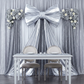 Silver metallic sweetheart table backdrop with a giant bow, flowing drapes, white table, and two chairs for a wedding.