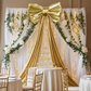 XL Giant Fabric Bow for Backdrop Metallic Gold against sheer white drapes panels, greenery garland and white florals behind a champagne table
