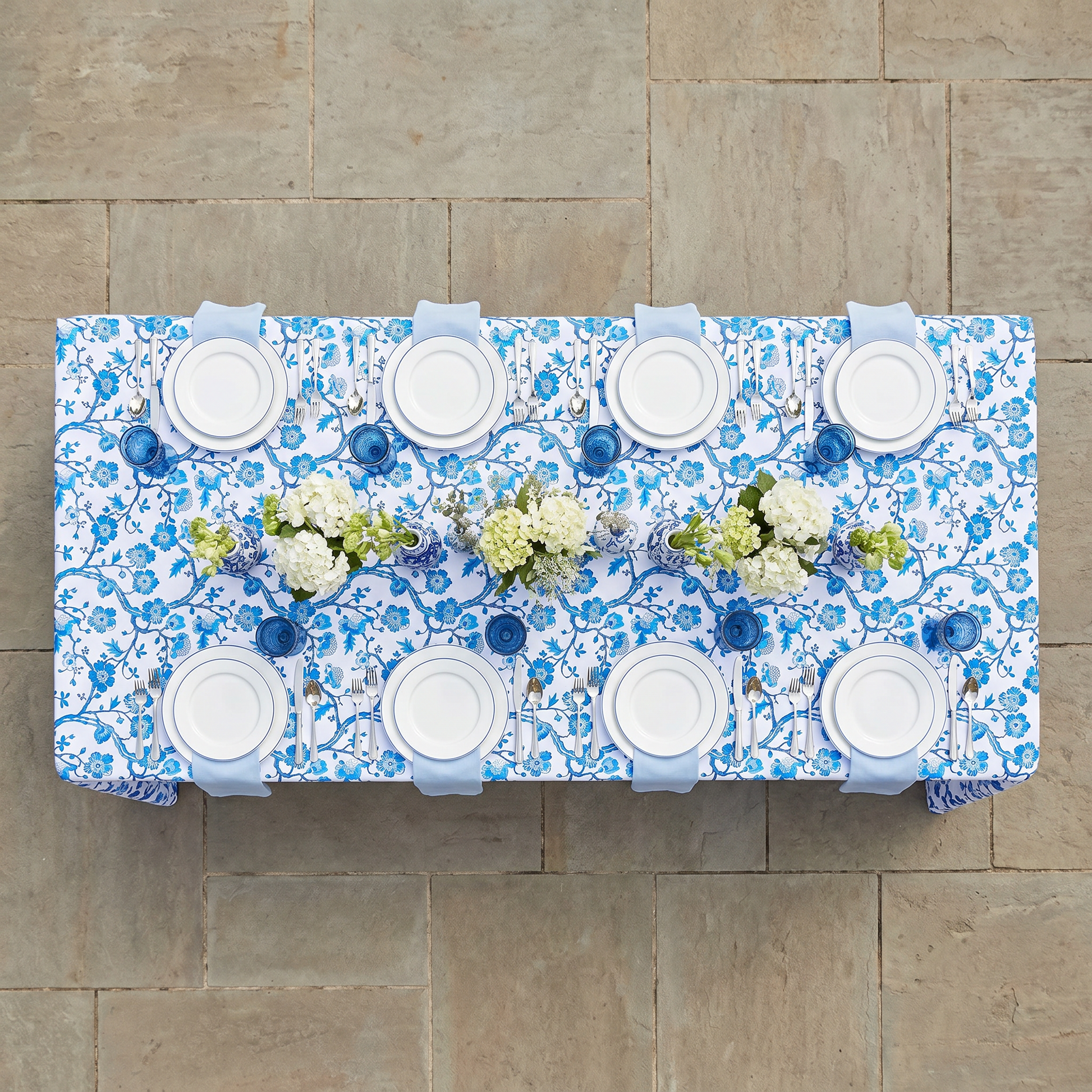 Chinoiserie blue rectangular tablecloth with floral pattern, top-down view, set with white plates and blue napkins.