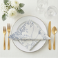 Bird's eye view lifestyle flat-lay of dusty blue leaf branch jacquard napkins on an elegant table setting with white plates, gold cutlery, and floral centerpiece