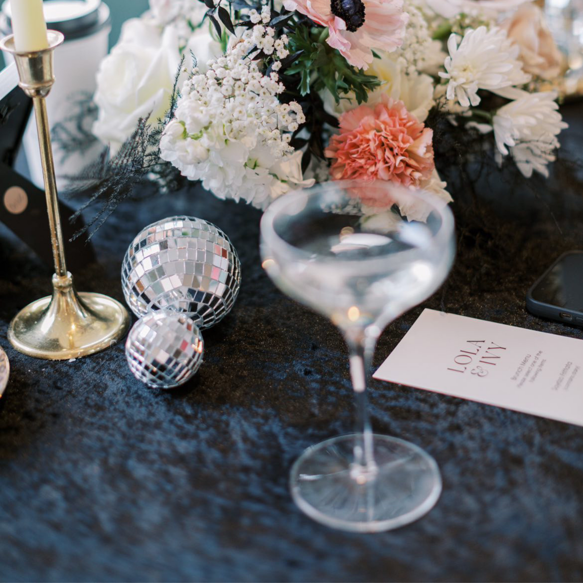 Decorative tablescape of black velvet tablecloth setup with flowers, disco balls, a cocktail glass, and a card.